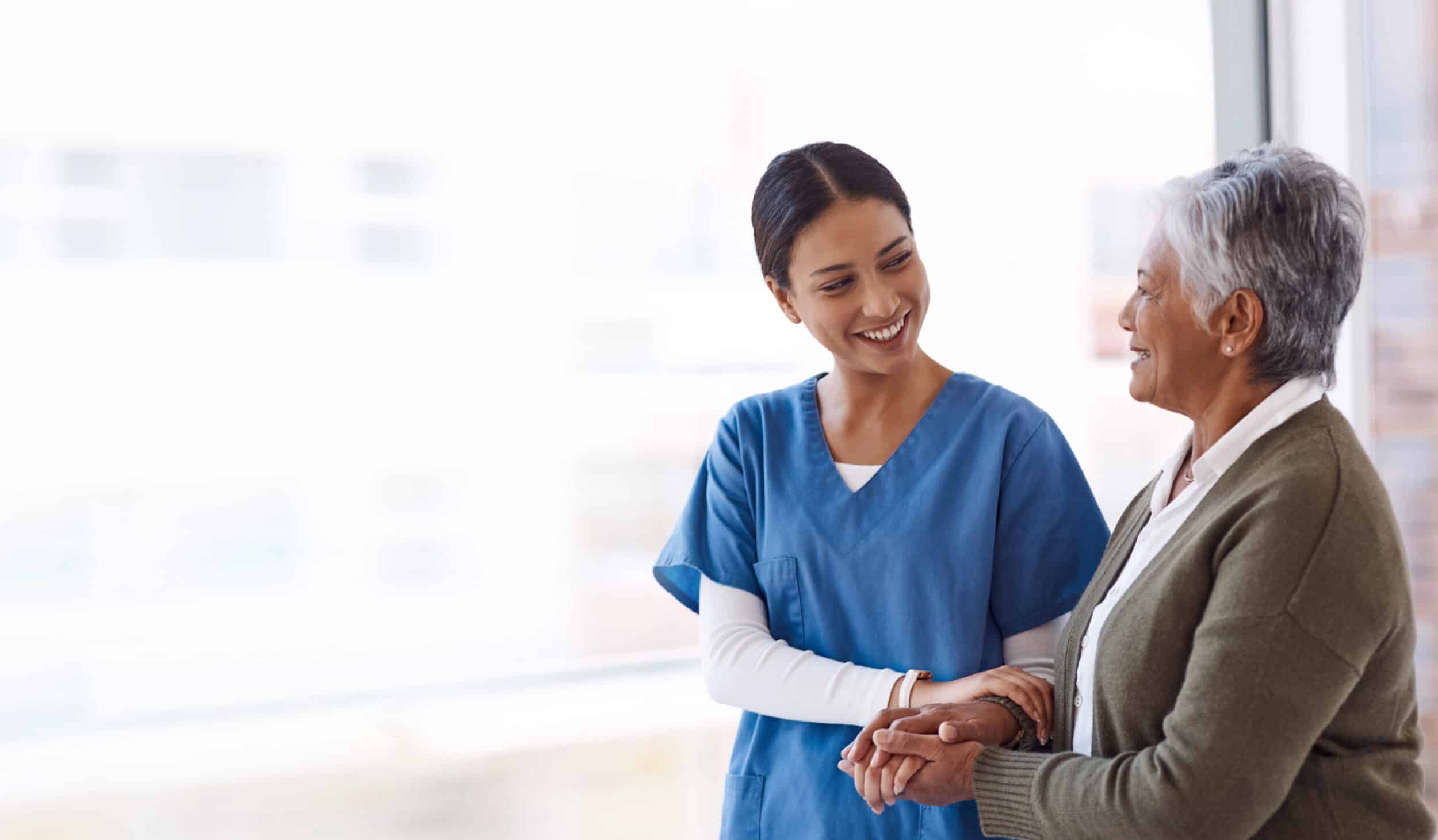 A healthcare professional in blue scrubs smiles while talking to an elderly woman in a green cardigan.