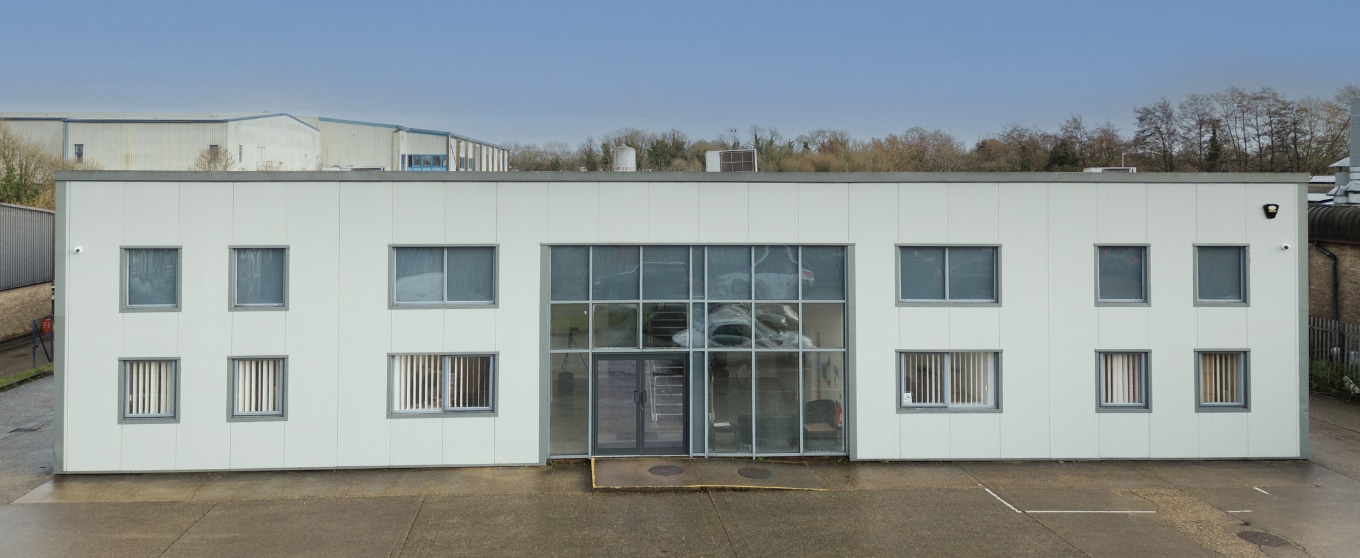 Contact us at our modern white industrial building with large glass entrance and multiple windows. Overcast sky above and wet ground in front.