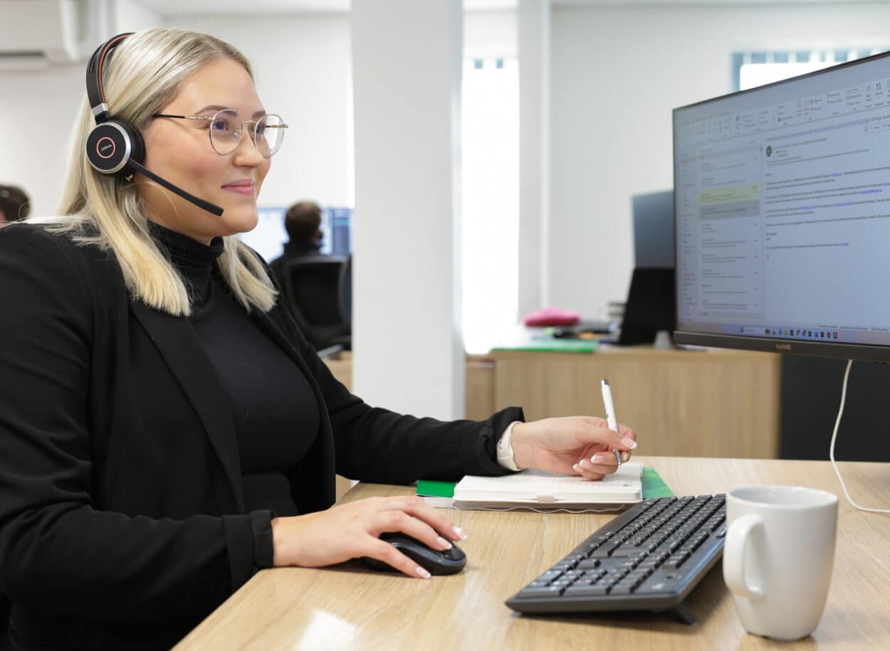 Woman wearing a headset works at a computer, holding a pen, with documents and a coffee mug on the desk. about