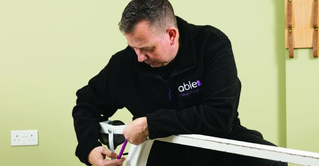 A man in a black fleece jacket is adjusting a component on a piece of furniture against a light green wall.