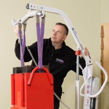 A person operates a patient lift device with slings attached to a red platform in a room with a beige wall. about