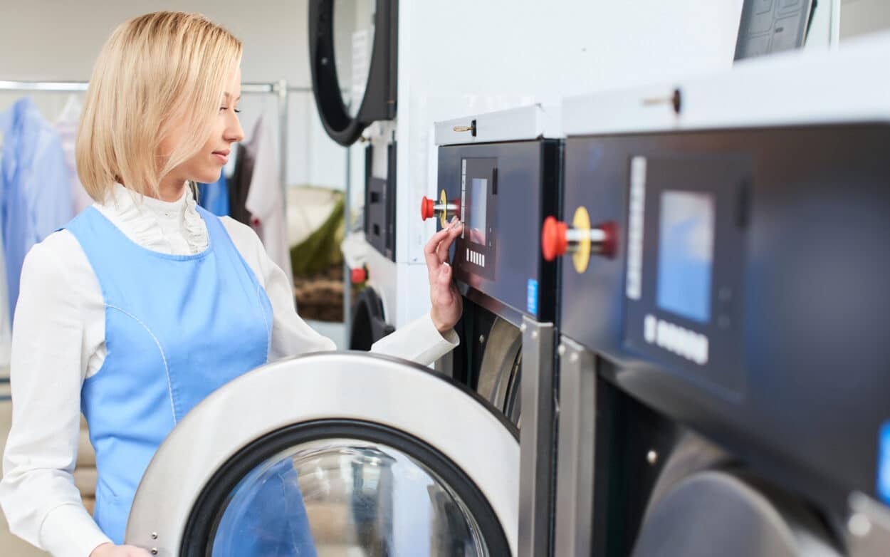 A woman operates a washing machine in a laundry facility, checking the auto dosing settings on the control panel.