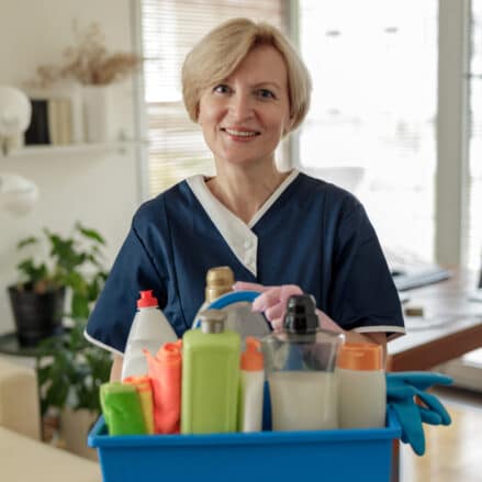 A person in a blue uniform holds a blue cleaning caddy filled with various cleaning supplies, including bottles, gloves, and cloths, standing in a room with plants and furniture. about