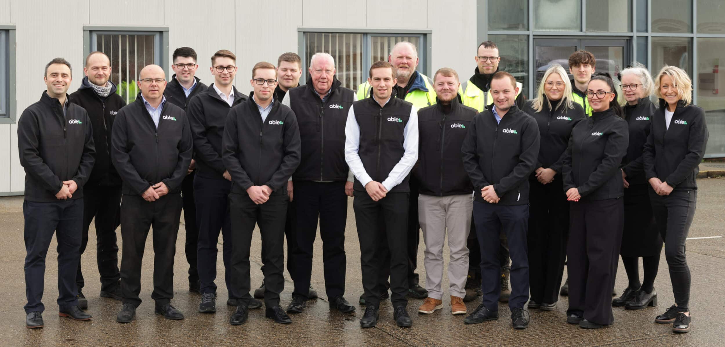 A group of 16 people standing in front of a building, wearing matching black jackets.