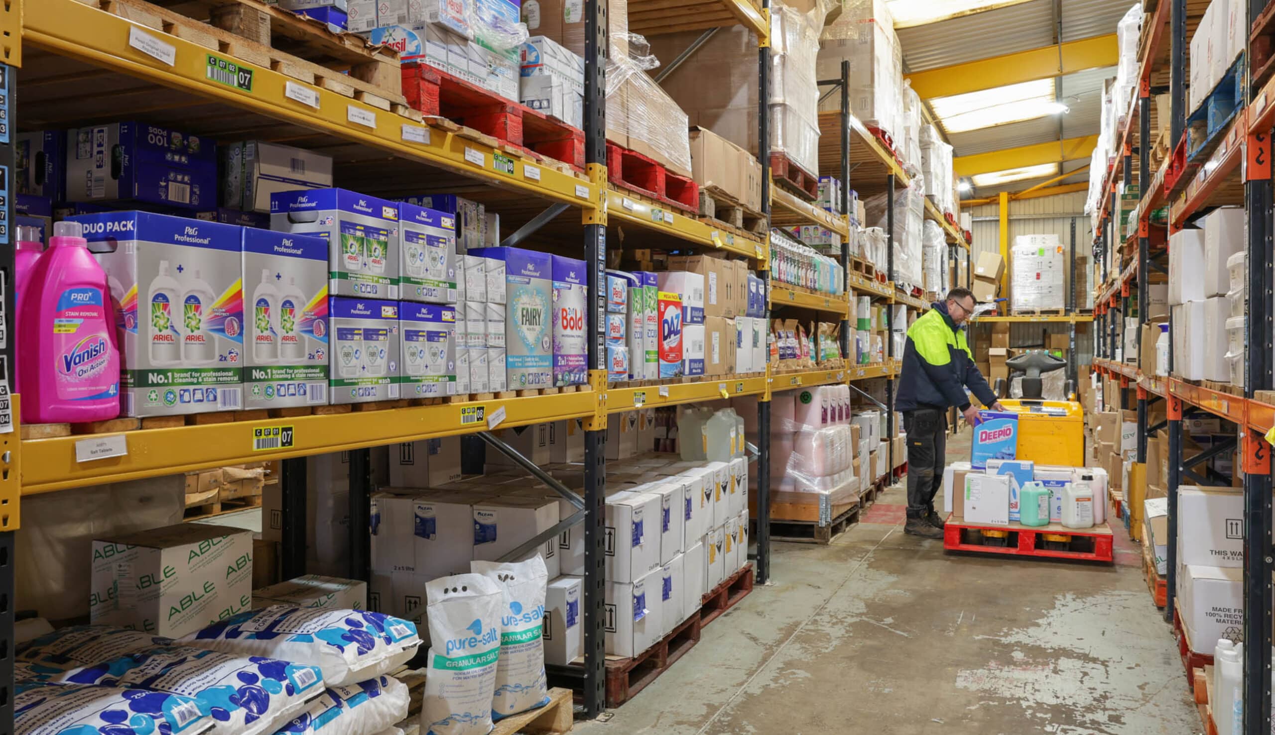 A person in a high-visibility jacket moves a cart through a warehouse aisle stocked with various household products on shelves.