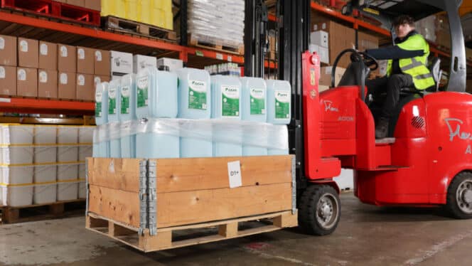 A person operates a red forklift, moving a wooden pallet with multiple large containers in a warehouse.