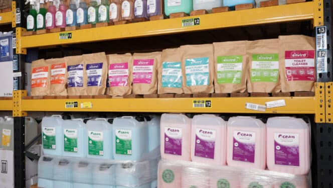 Shelves stocked with eco-friendly cleaning products in various packages and colors. Cardboard boxes and plastic containers labeled with brands are organized on metal shelving.