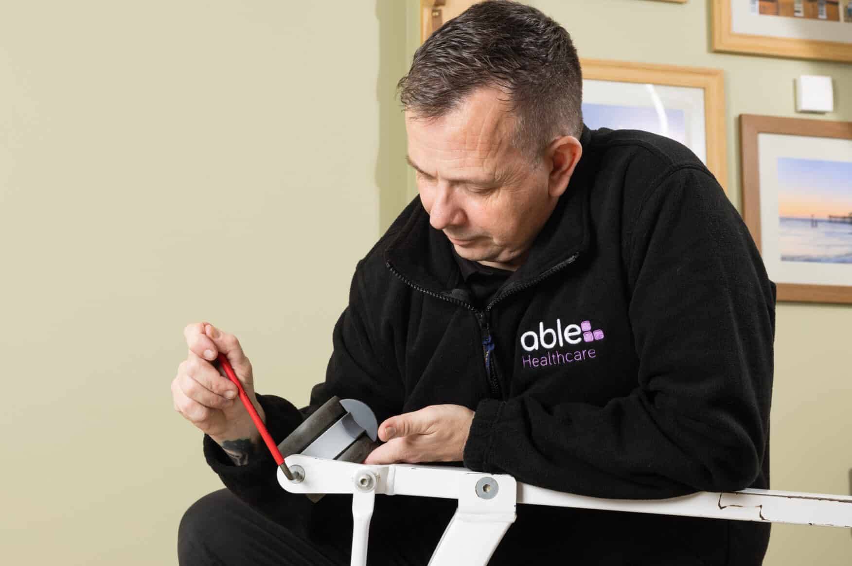 A man in a black jacket is servicing a care home hoist with a red brush, seated indoors by a wall with framed pictures as part of their care home equipment servicing.