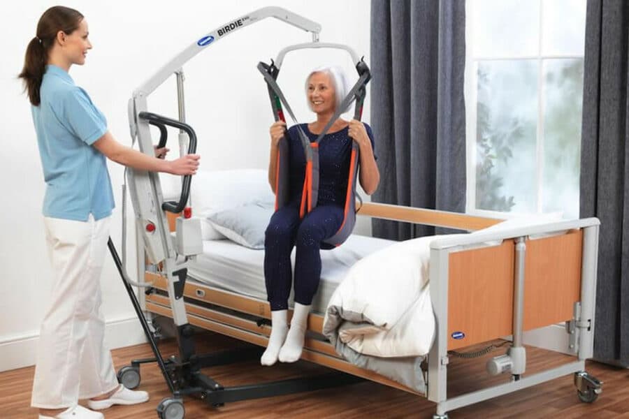 A caregiver operates a patient lift to assist a woman sitting in a sling over a hospital bed in a bright room with wooden flooring.