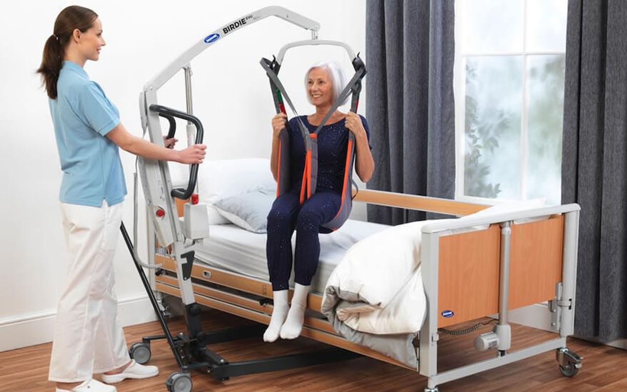 A caregiver operates a patient lift to assist a woman sitting in a sling over a hospital bed in a bright room with wooden flooring.