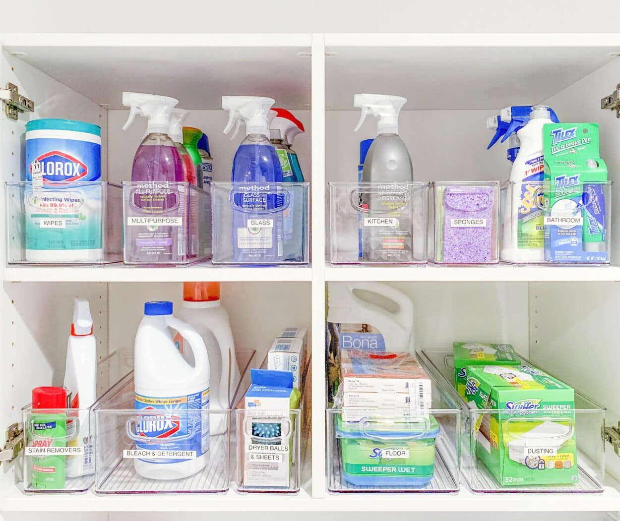 Neatly organized white cabinet with clear bins containing various household cleaning supplies, sprays, wipes, detergents, and sponges.