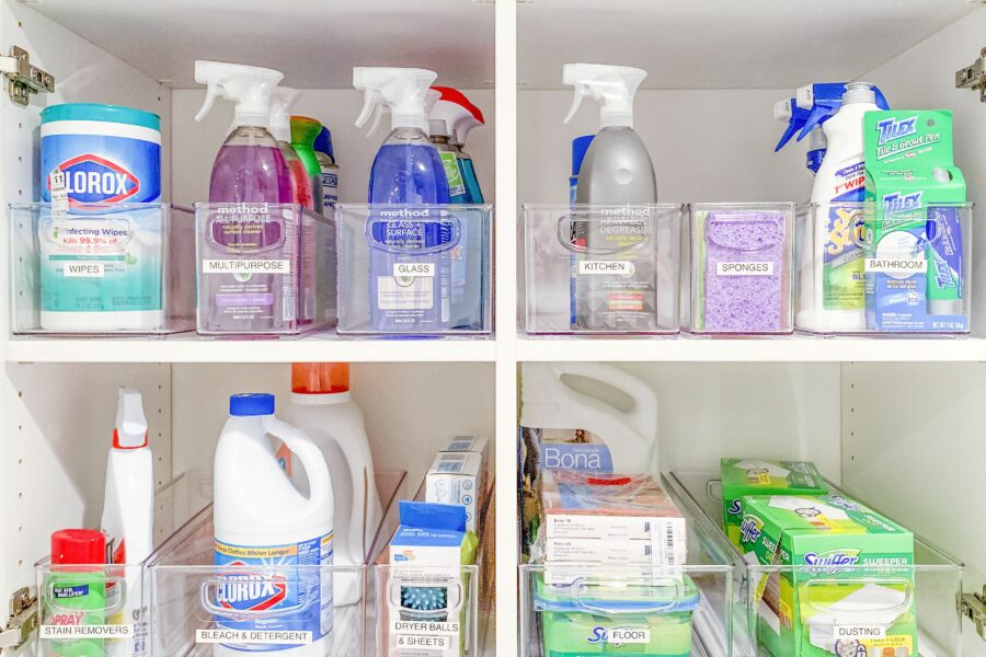 Neatly organized white cabinet with clear bins containing various household cleaning supplies, sprays, wipes, detergents, and sponges.