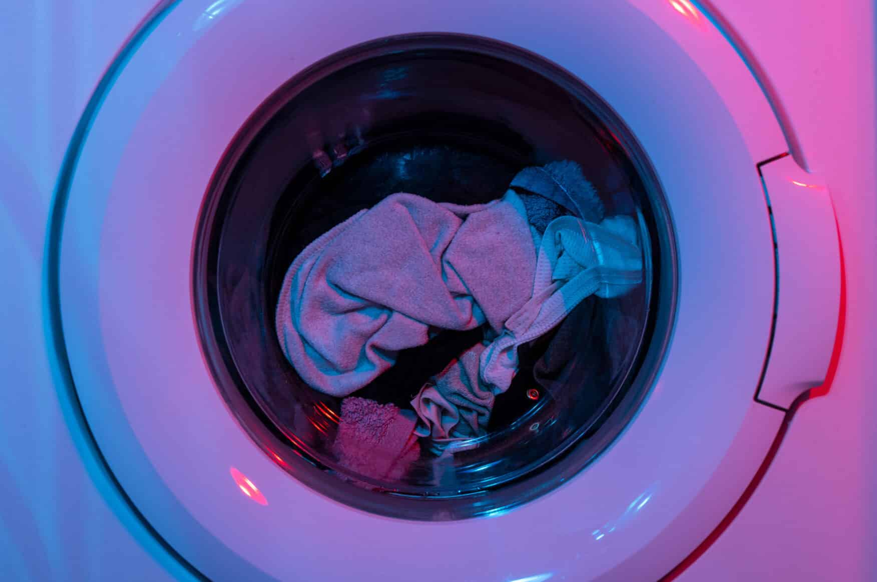 A front-loading washing machine with a few pieces of clothing inside, photographed under blue and pink lighting, highlights the importance of using care home laundry chemicals for effective cleaning and hygiene.