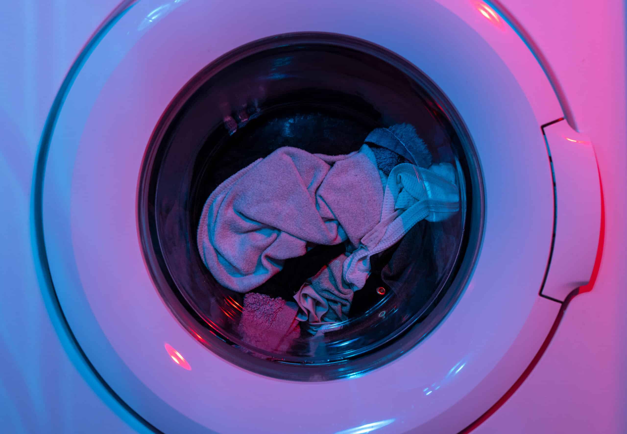 A front-loading washing machine with a few pieces of clothing inside, photographed under blue and pink lighting, highlights the importance of using care home laundry chemicals for effective cleaning and hygiene.