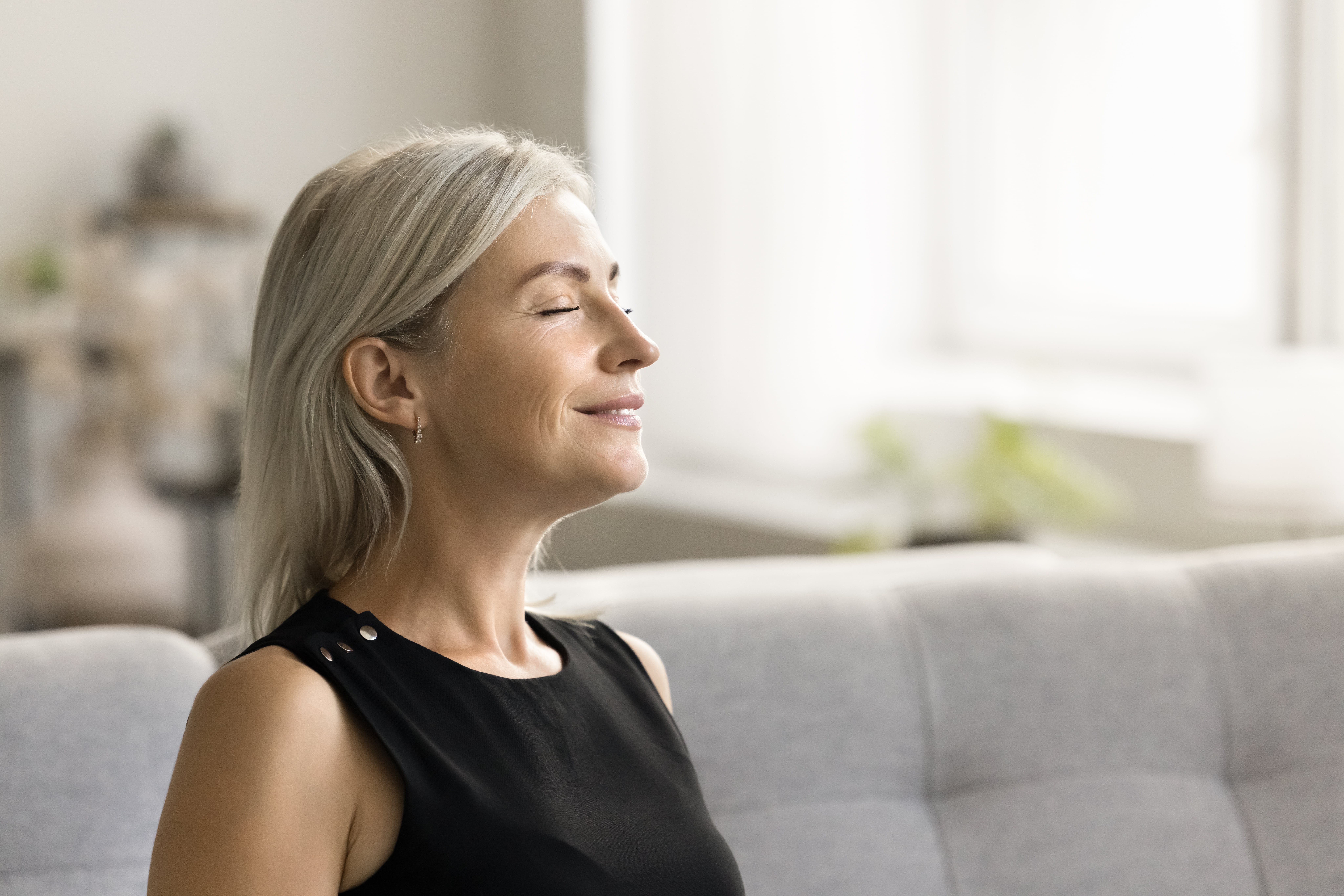 A woman with gray hair sits on a couch, eyes closed, wearing a sleeveless black top, appearing calm and relaxed in a bright room designed for natural odour control care homes.
