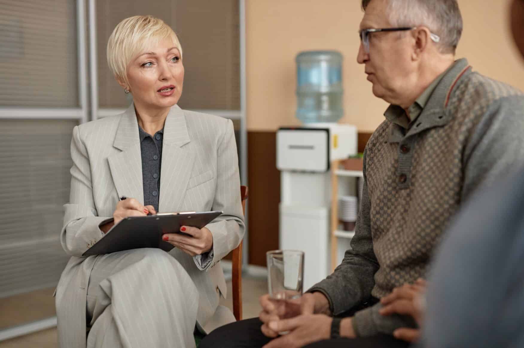 A woman in a suit holds a clipboard and discusses a mock CQC Audit with a seated man holding a glass of water in an office setting, with a water cooler visible in the background./ They are preparing via a Mock CQC audit