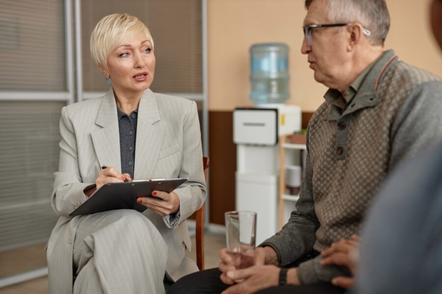 A woman in a suit holds a clipboard and discusses a mock CQC Audit with a seated man holding a glass of water in an office setting, with a water cooler visible in the background./ They are preparing via a Mock CQC audit