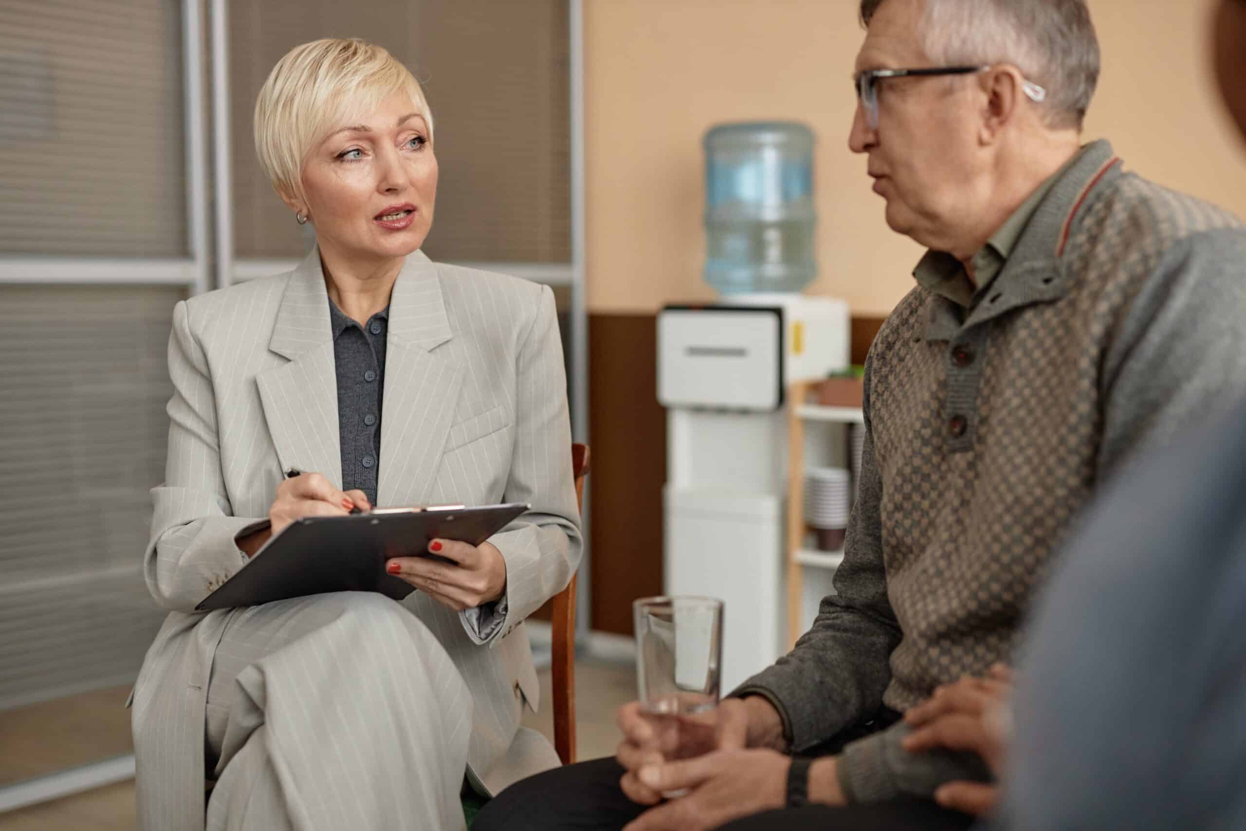 A woman in a suit holds a clipboard and discusses a mock CQC Audit with a seated man holding a glass of water in an office setting, with a water cooler visible in the background./ They are preparing via a Mock CQC audit