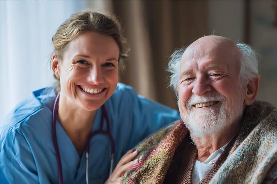 A nurse in scrubs smiles beside an elderly man wrapped in a blanket, both looking at the camera in a well-lit room.