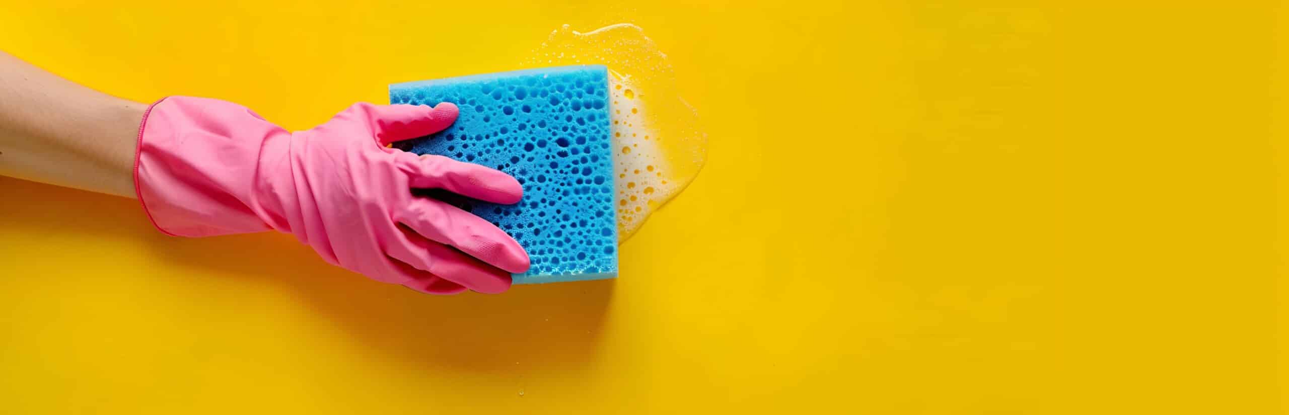 Hand wearing a pink glove cleaning a yellow surface with a blue sponge as part of care home cleaning tasks.